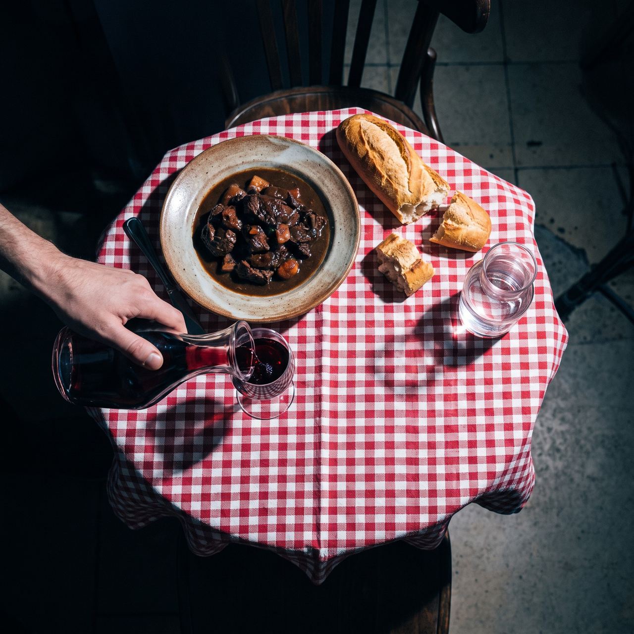 Vue de dessus d’une table de bistrot : nappe à carreaux, plat mijoté, main qui verse du vin.
